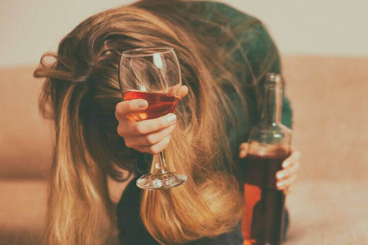 Frau mit nach unten geneigtem Kopf, die ein Weinglas hält und traurig oder müde wirkt, mit einer Flasche neben sich auf einem Sofa, in einer dunklen, häuslichen Atmosphäre.