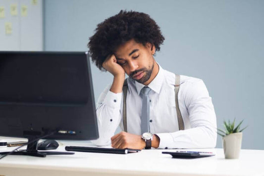 Müder Geschäftsmann, der seinen Kopf mit der Hand stützt, sitzt an seinem Arbeitsplatz mit einem ausgeschalteten Computerbildschirm und Büromaterial in einem hellen, modernen Büro.