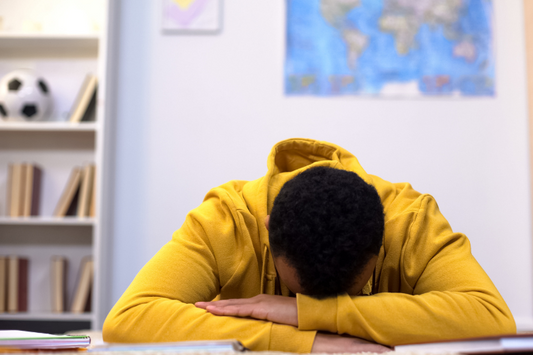 Un étudiant, assise à un bureau avec la tête reposant sur ses bras croisés, suggérant un état de fatigue ou d'épuisement.