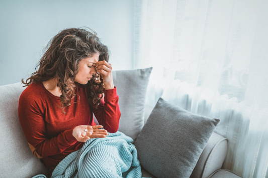 femme assise sur un canapé, manifestant des signes de douleur ou d'inconfort, probablement une migraine. Elle a une main sur son front et tient des médicaments dans l'autre main.