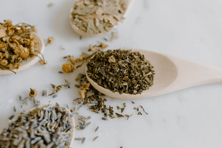 Trois cuillères en bois sur une surface blanche contenant différentes herbes séchées : menthe, camomille et lavande.