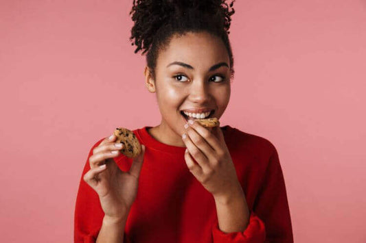 Jeune femme souriante aux cheveux bouclés en pull rouge mangeant un cookie aux pépites de chocolat sur un fond rose uni.