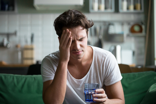  Un homme semble souffrir d'un mal de tête, tenant sa tête avec une main et un verre d'eau dans l'autre. Cette image peut illustrer des conseils pour éviter la gueule de bois.