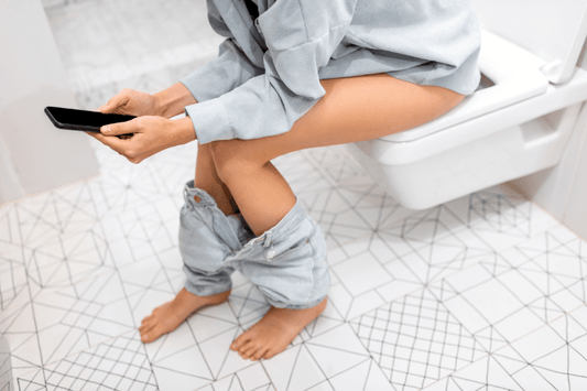 Femme assise sur des toiletttes qui à son pantaon baissé et qui est sur son smartphone en attendant de finir ses besoins